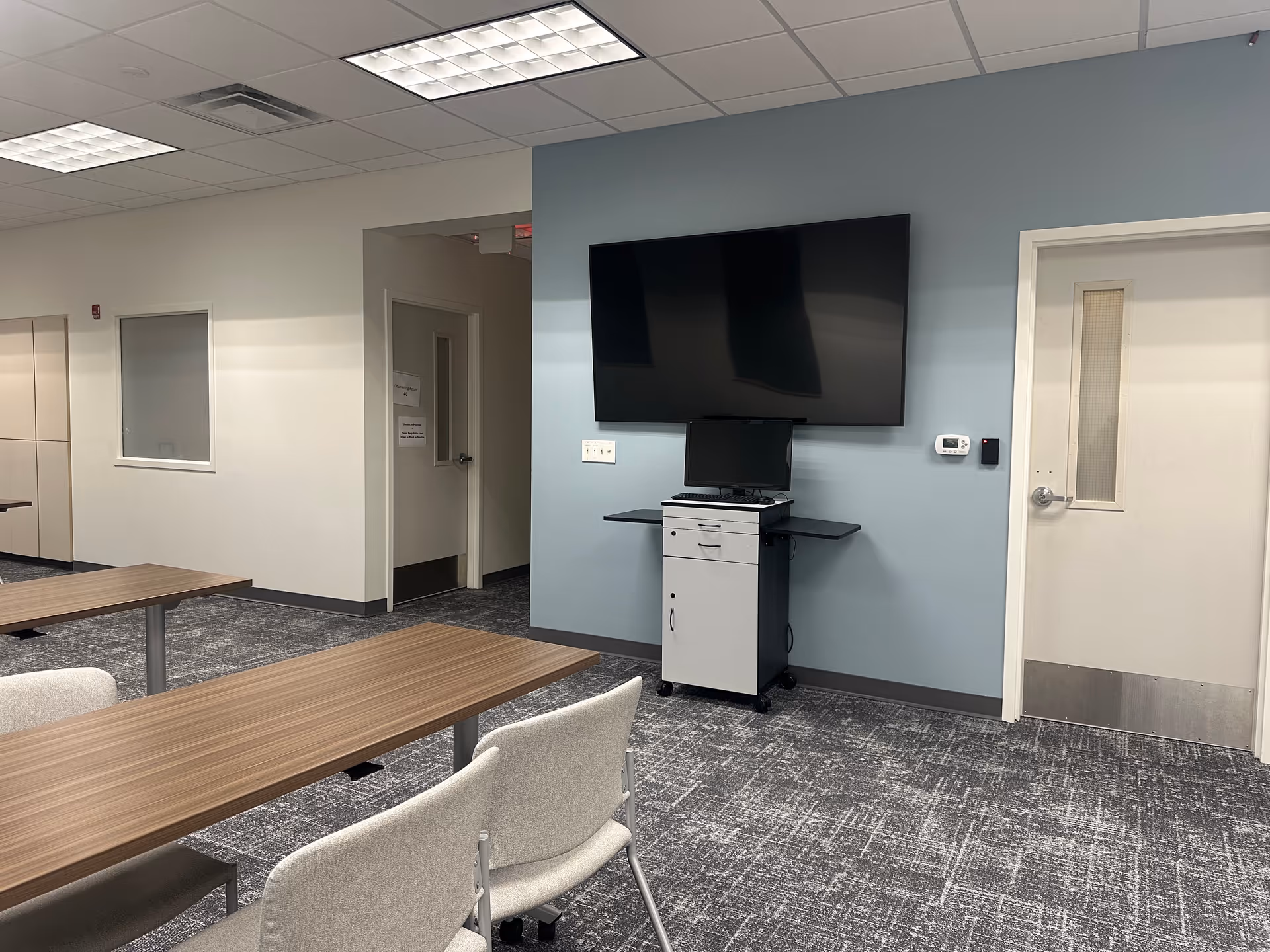 Conference room with tables, chairs, a wall-mounted TV and a cart with computer against blue wall.
