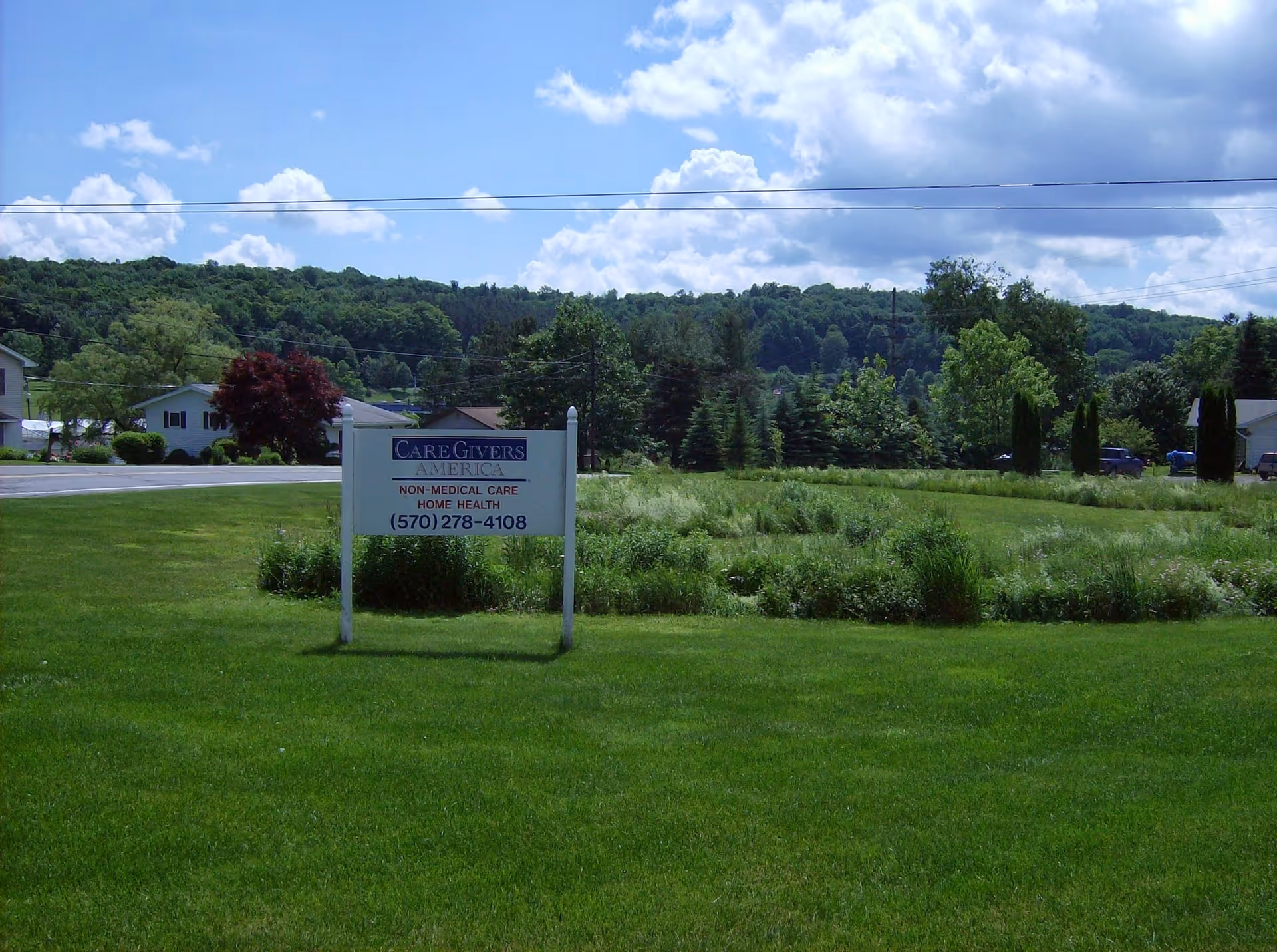 Wide grassy lot with CareGivers America sign and distant trees under blue sky