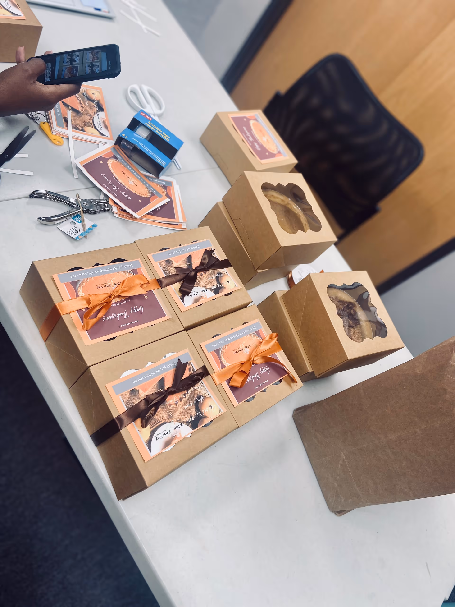 Desk with ribbon-wrapped gift boxes, cards, scissors, and a person using a smartphone.