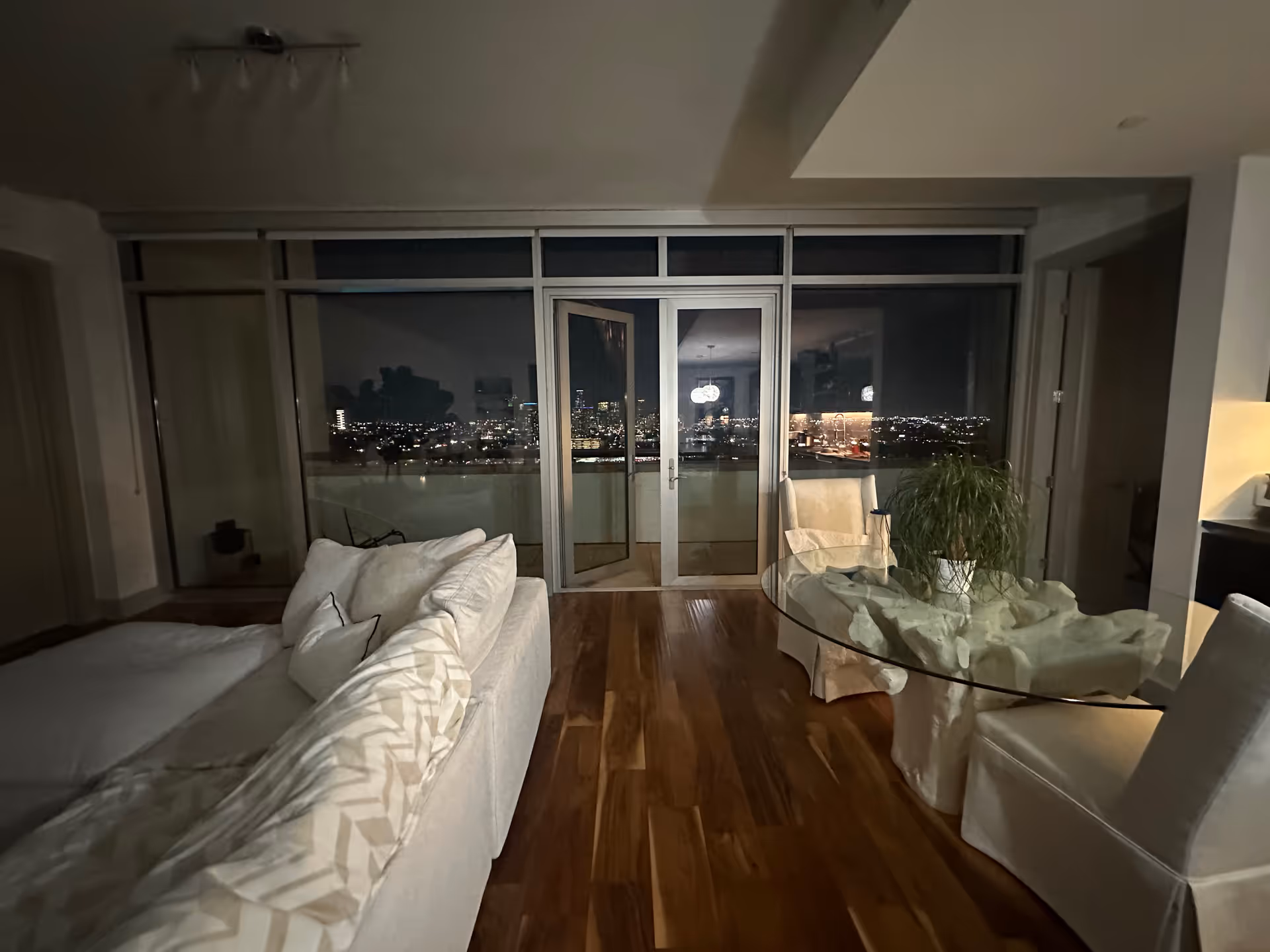 Modern living room with white furniture, glass round table, plant, and city view through balcony doors.