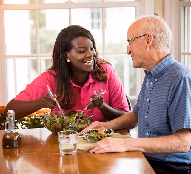 A caregiver and elderly man share a meal at a sunlit dining table.