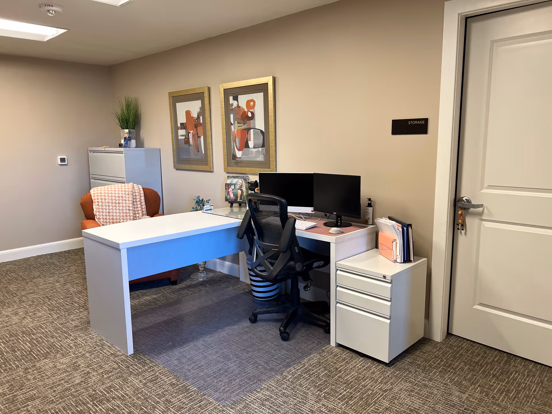 Office workspace with desk, chair, dual monitors, filing cabinet, and storage sign.