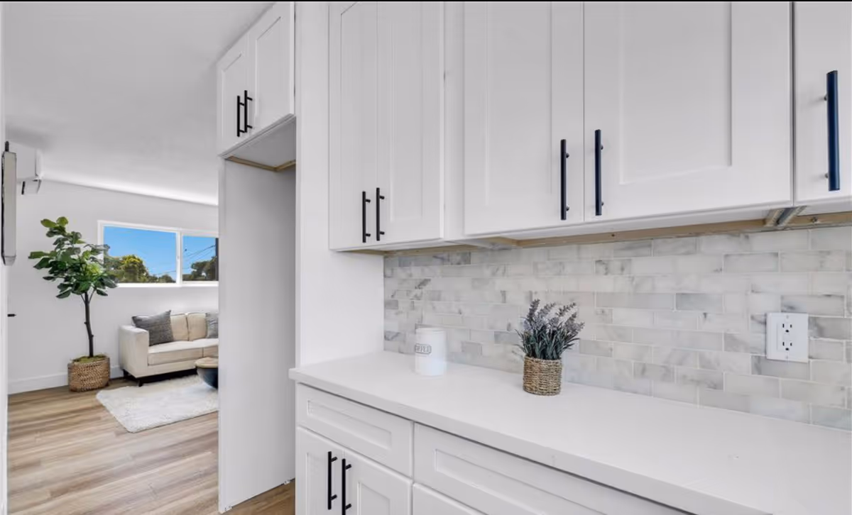 Bright modern kitchen with white cabinets, light backsplash, opening to a living area.