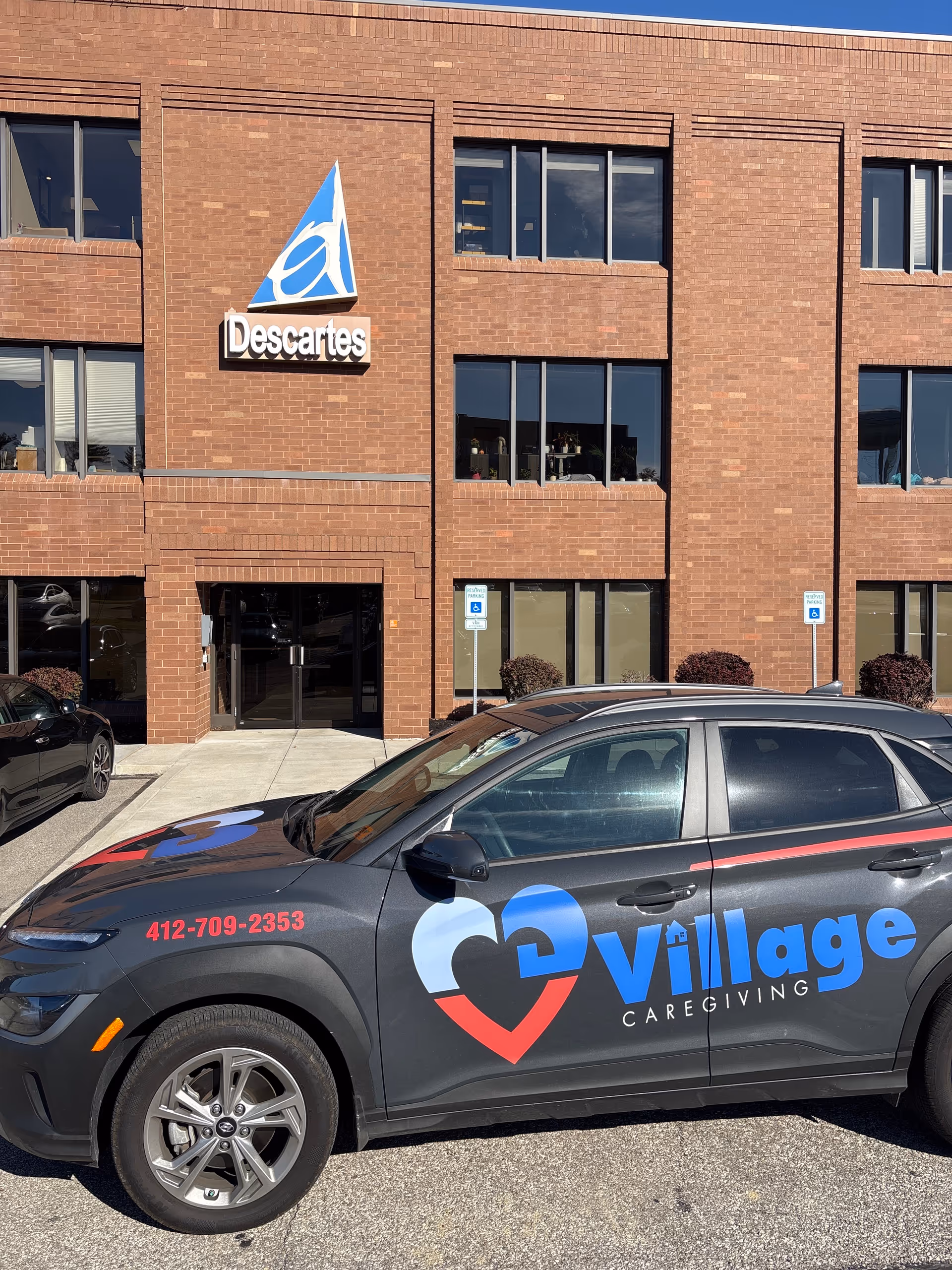 Brick building exterior with a Descartes sign and Village Caregiving vehicle parked in front.