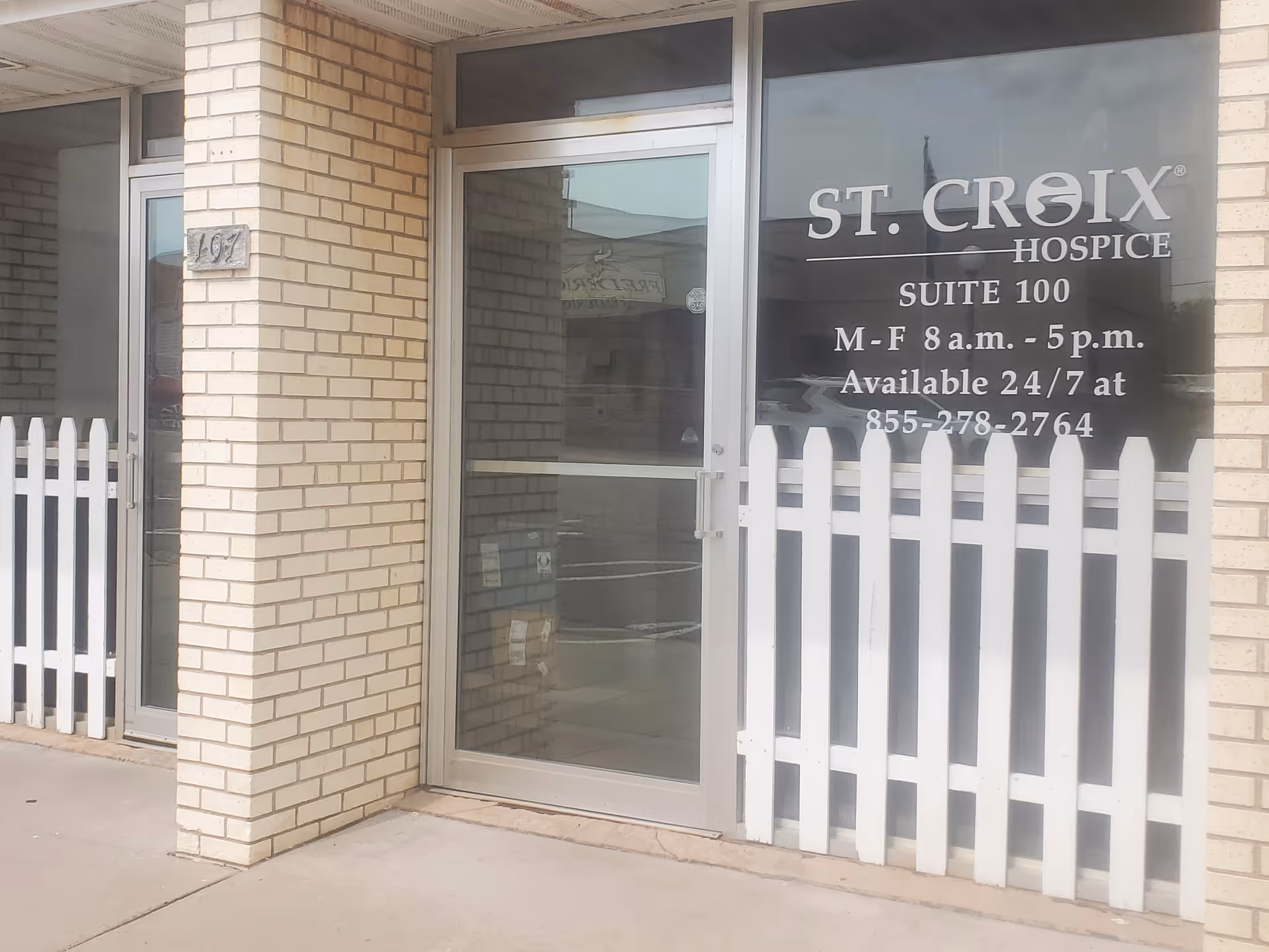 Front entrance of St. Croix Hospice with glass door, brick pillars, and white picket fence.