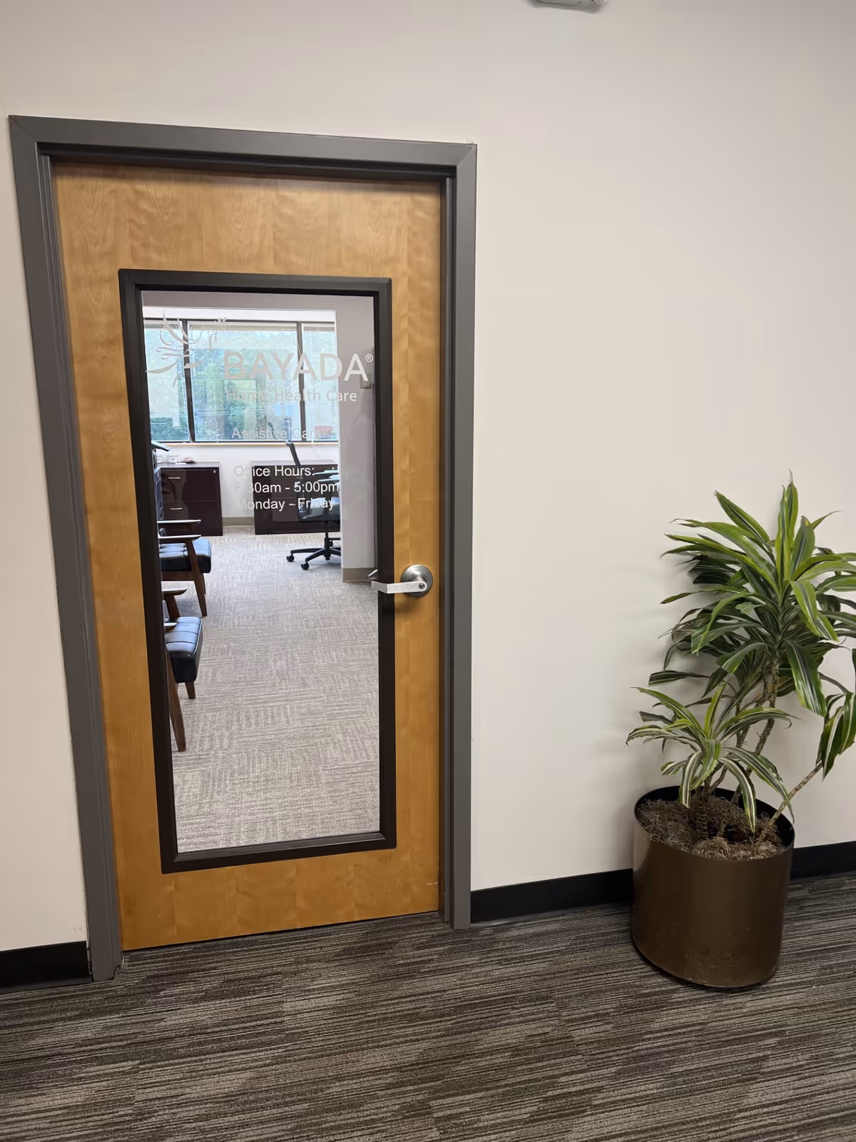 Wooden interior door with glass panel displaying BAYADA Assistive Care, office furniture visible beyond, plant nearby.