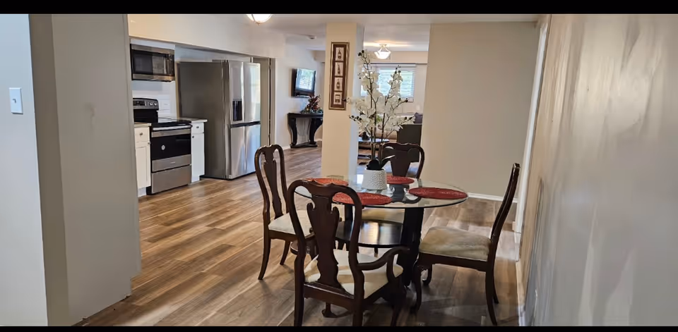 Dining table with four chairs in a senior living facility dining area; kitchen visible in background.