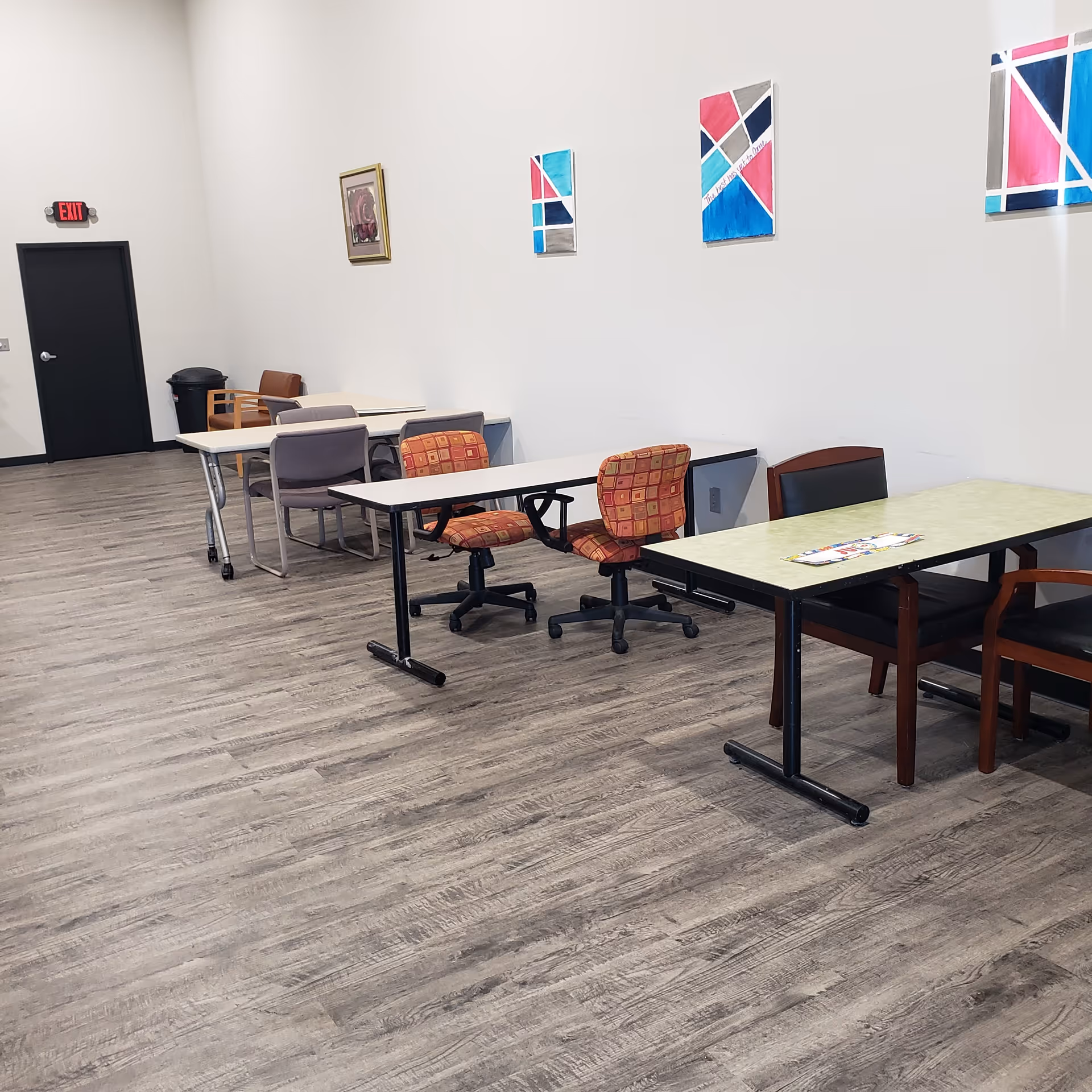 A bright, empty common room with several tables, chairs, wall art, and an exit door.