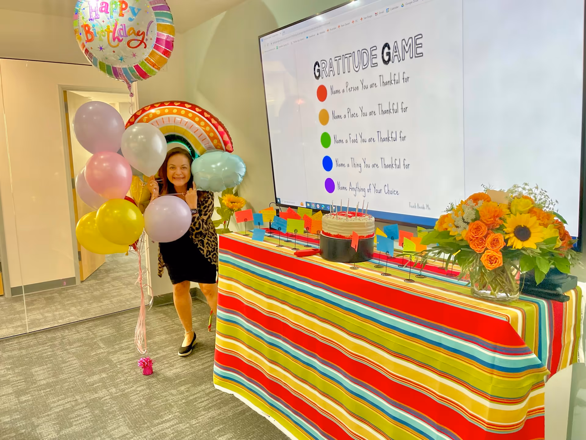 Celebration setup with balloons, cake, flowers, and a staff member near a striped table.