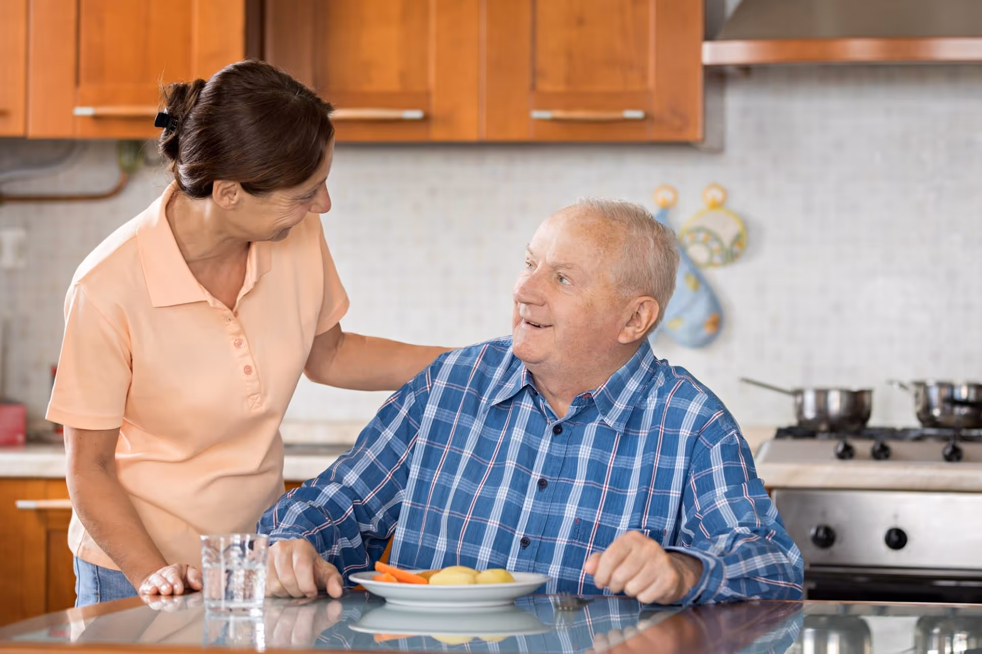 Caregiver chats with an elderly man at a kitchen island, plate of fruit and glass nearby.