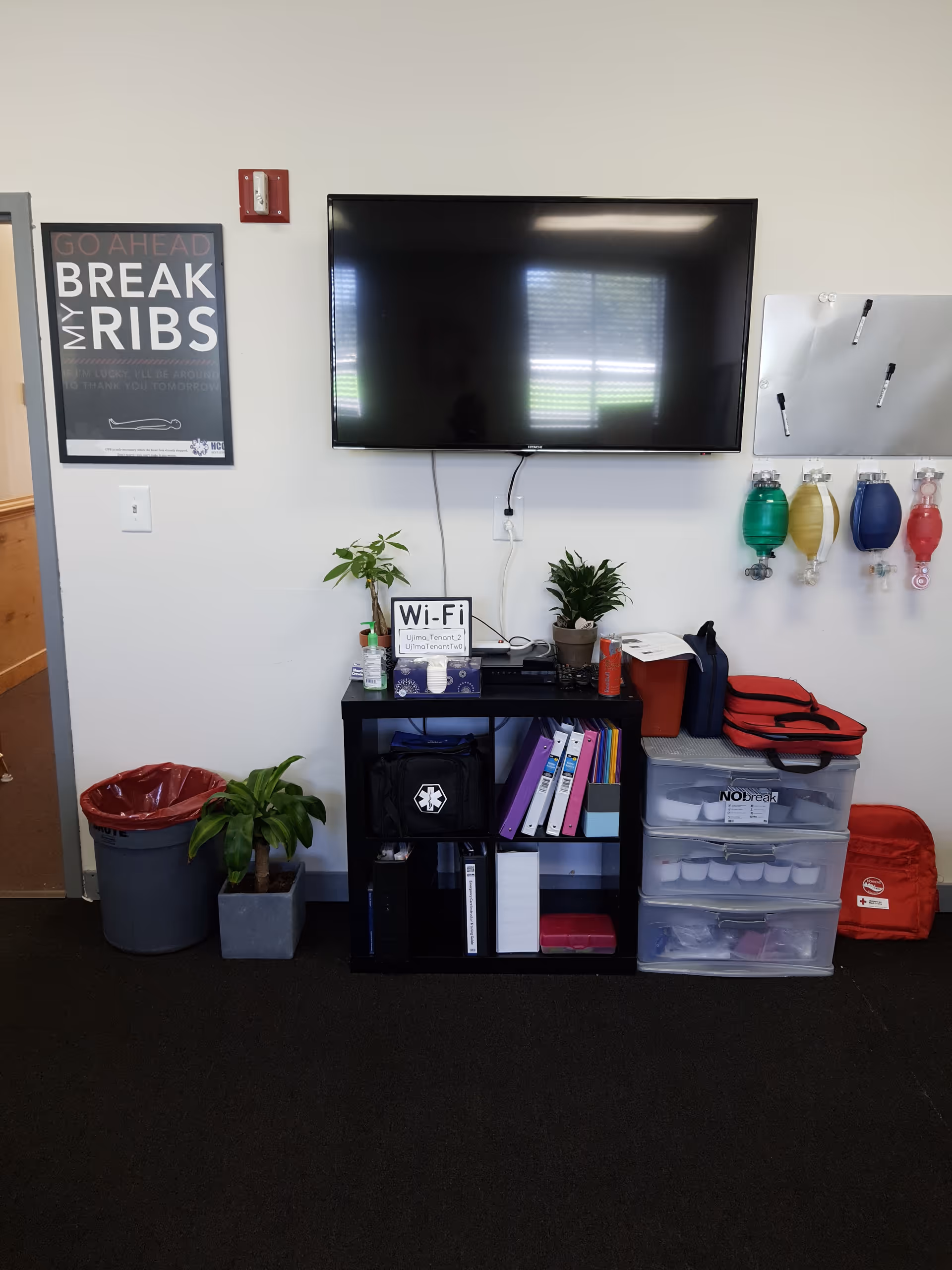 Mounted TV above a shelf with plants, binders, and storage drawers in a facility common area.