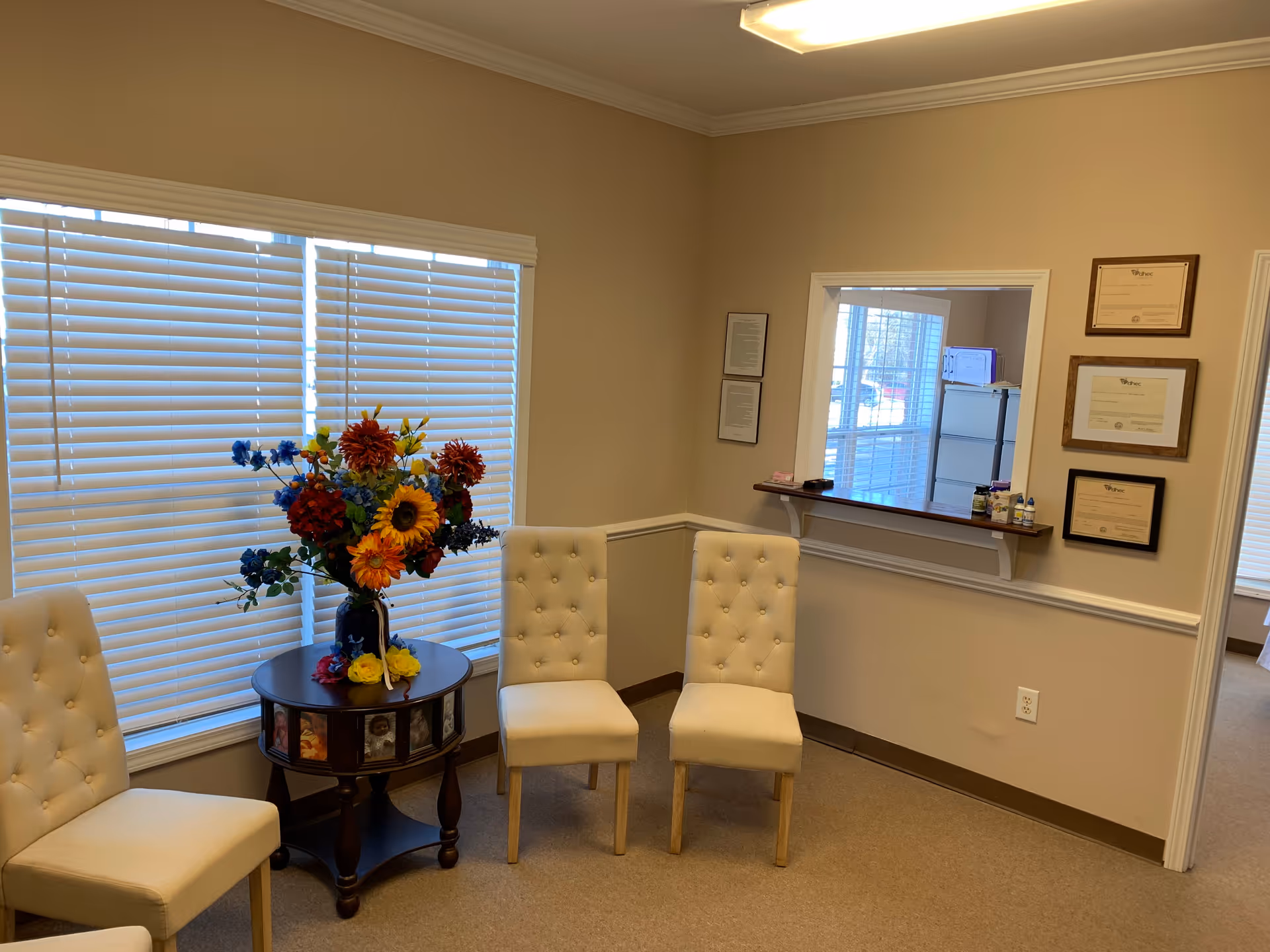 Lobby seating area with four cream tufted chairs, a floral arrangement, blinds, and certificates.