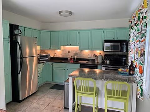 Mint-green kitchen with stainless appliances, island seating, and colorful patterned curtains.