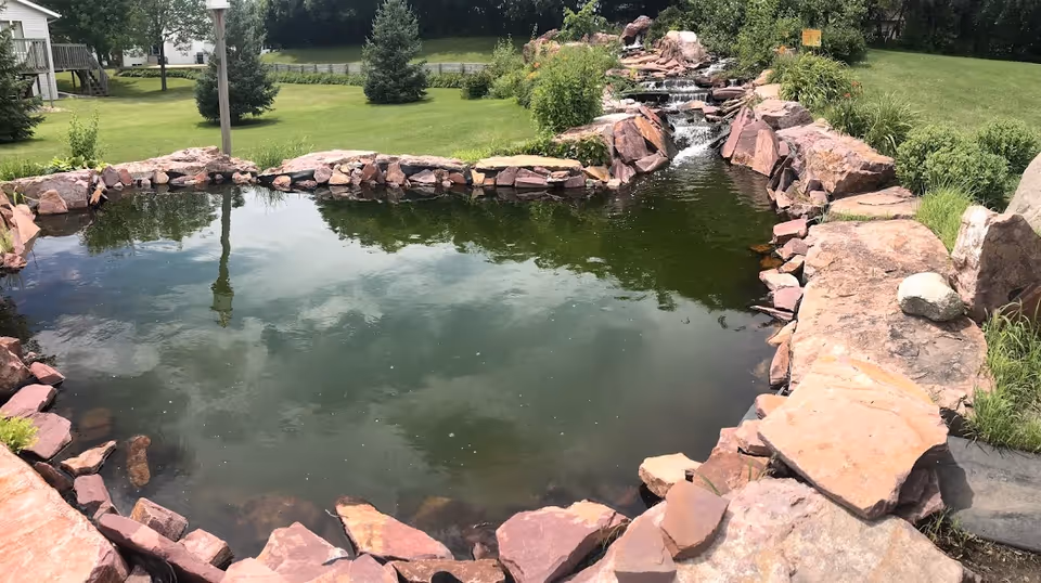Stone-edged pond with a waterfall on a grassy hospice campus.