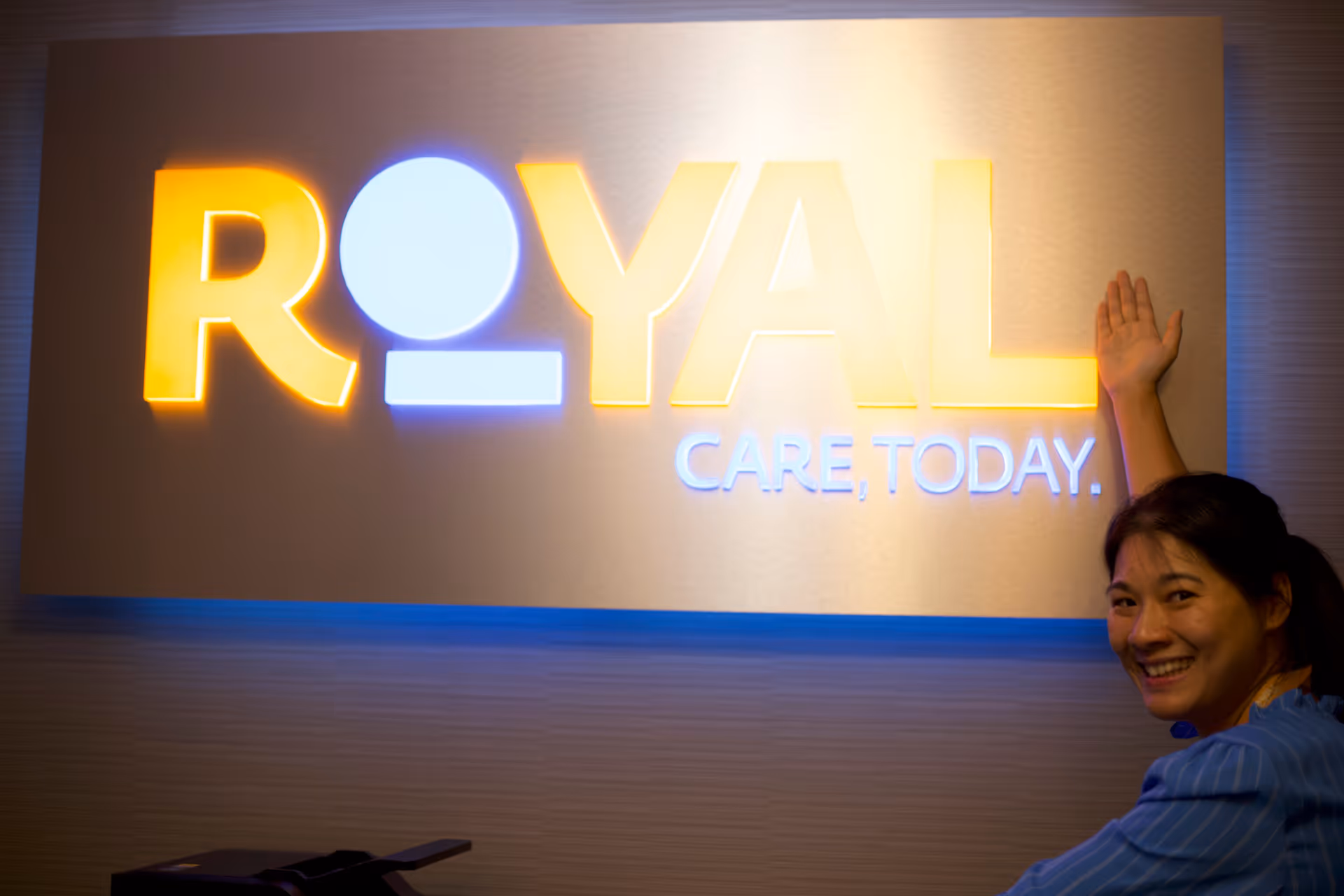 Bright illuminated ROYAL CARE sign on a wall with a smiling attendant beside it.