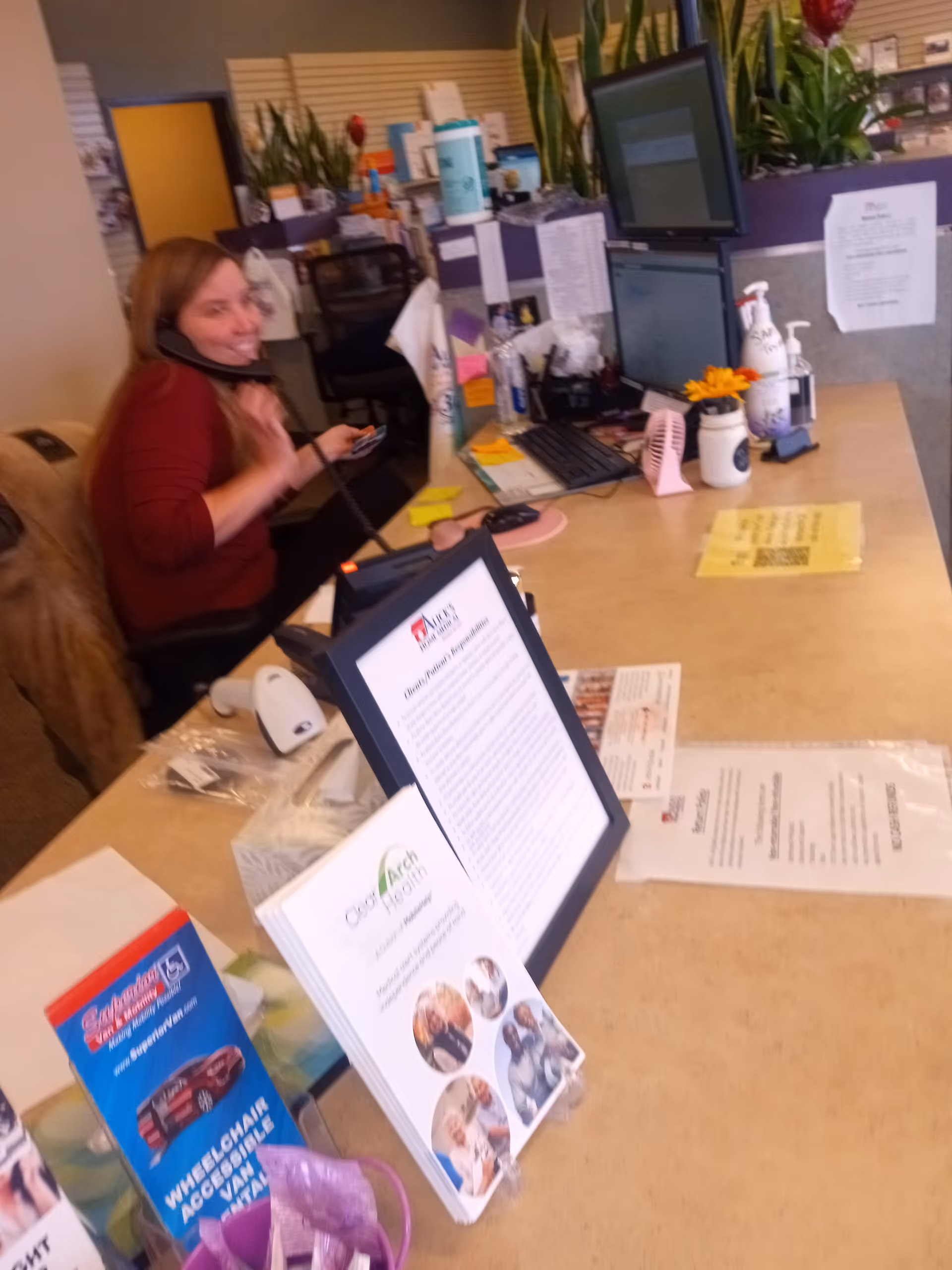 Receptionist seated at a front desk with computers and office supplies inside a medical facility.