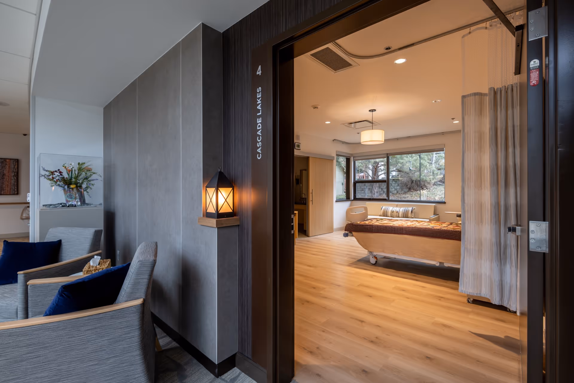 An open doorway reveals a patient room with a bed, curtain, and a seating lounge nearby.