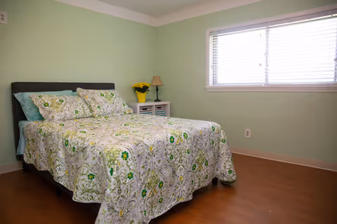 A simple bedroom with a queen bed, nightstand, and window in pastel green walls.