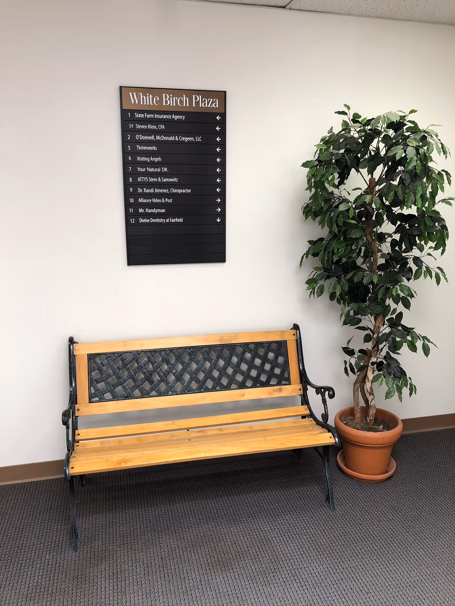 Wooden bench beside a potted plant and wall directory sign in a senior living hallway.