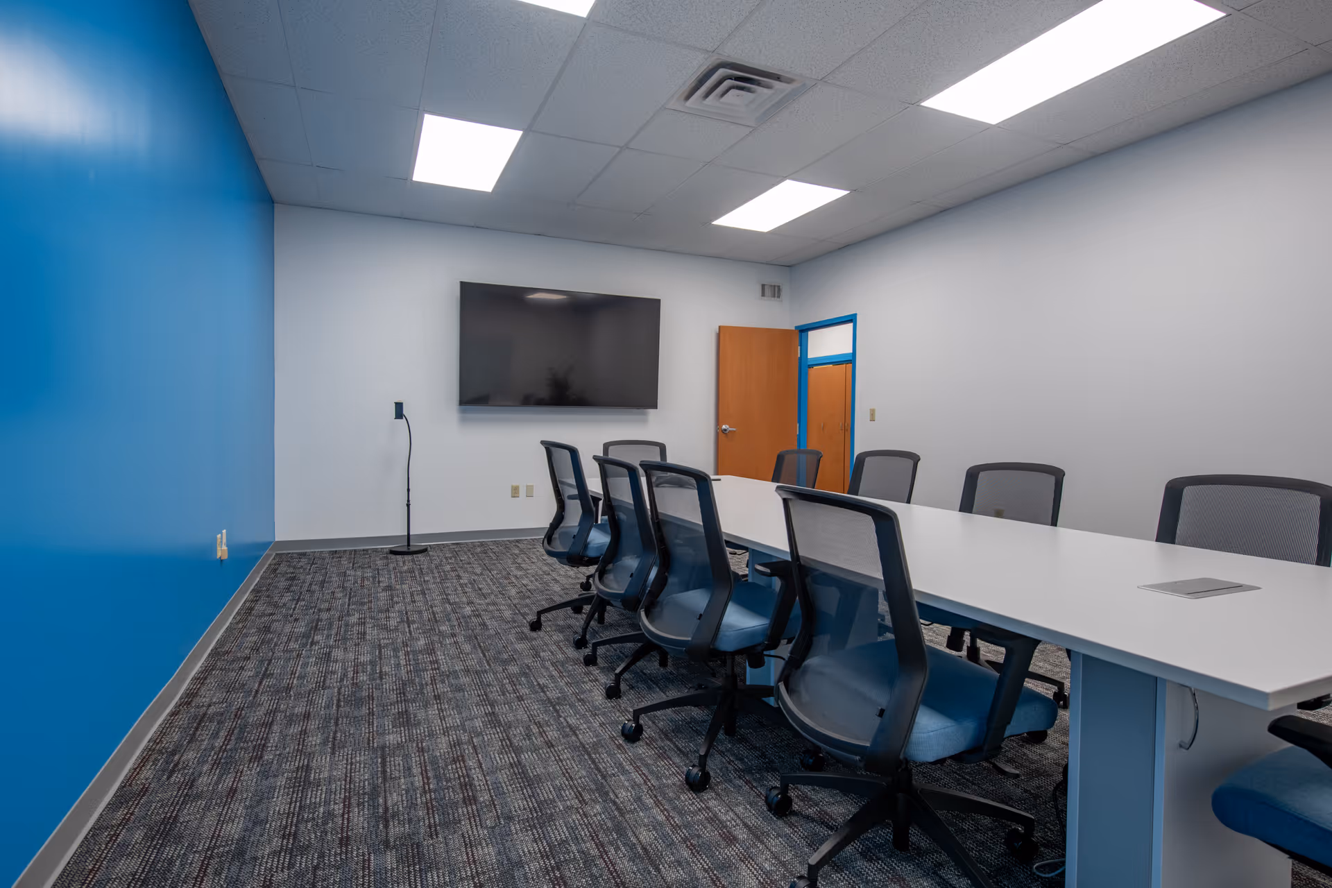 A modern conference room with a long white table, blue chairs, and a wall-mounted TV.