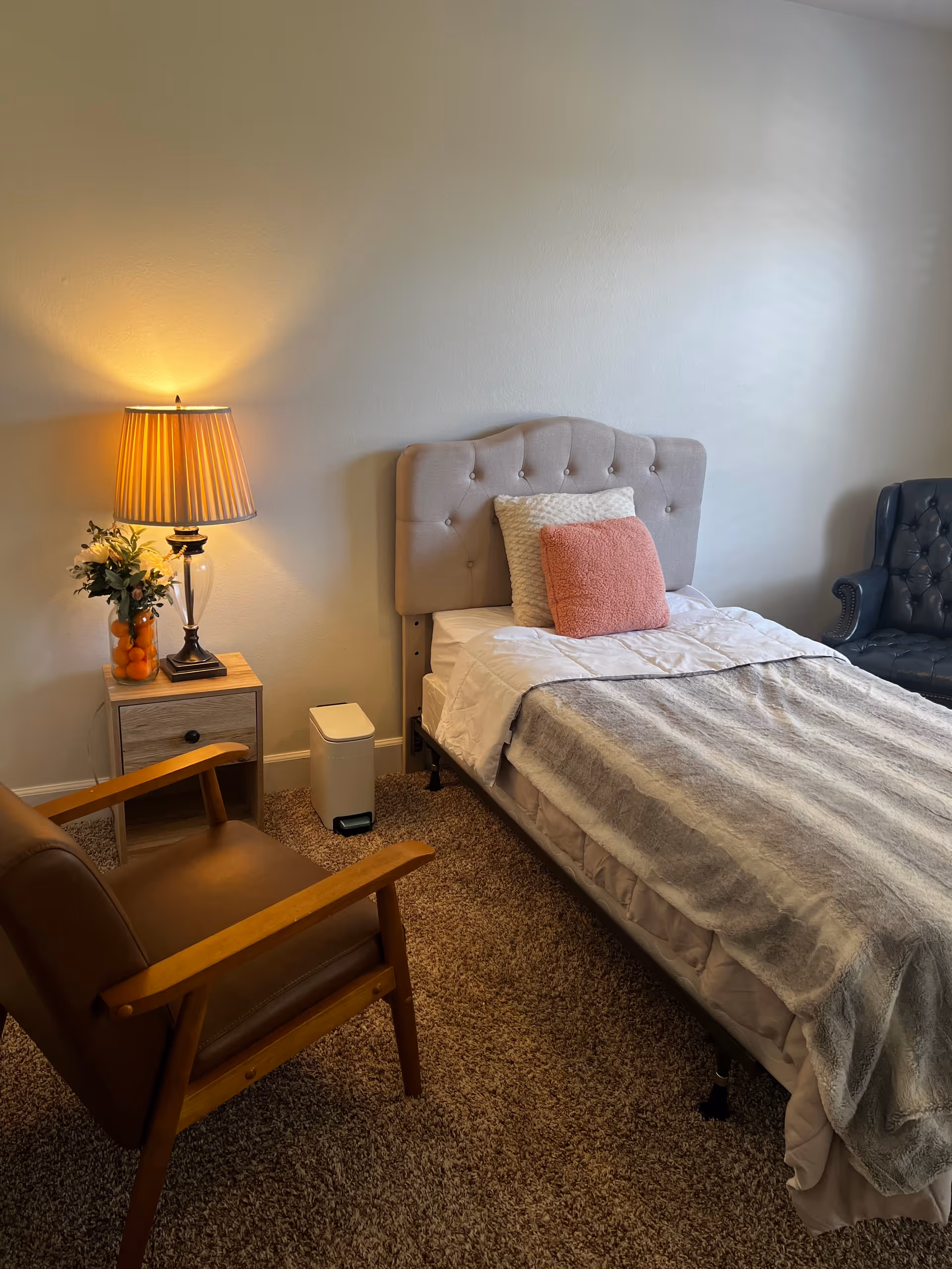 A cozy bedroom with tufted headboard, lamp, nightstand, and armchair in a care facility.