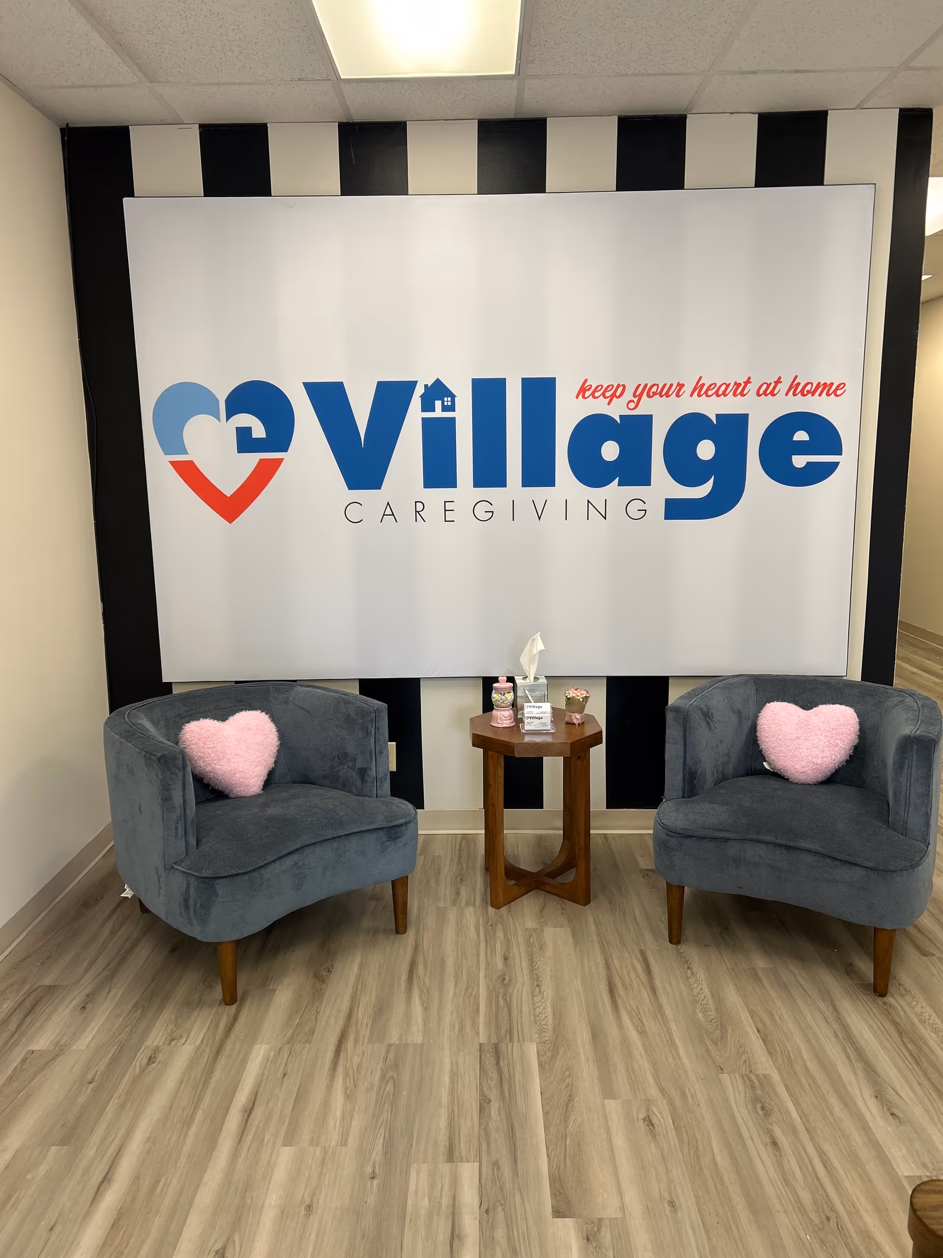 Two blue armchairs with pink heart pillows flank a small table under a Village Caregiving sign.