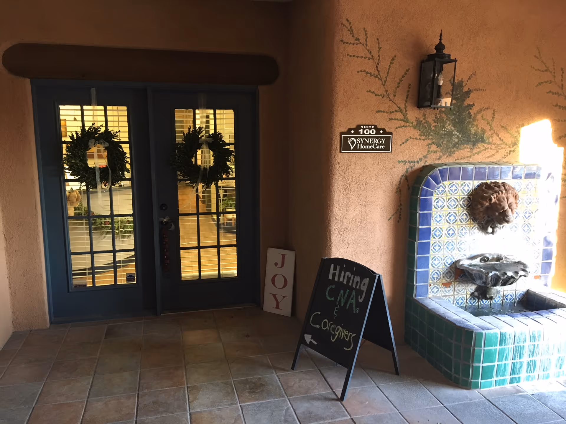 Blue double doors with wreaths flank tiled fountain outside a Synergy HomeCare entrance.