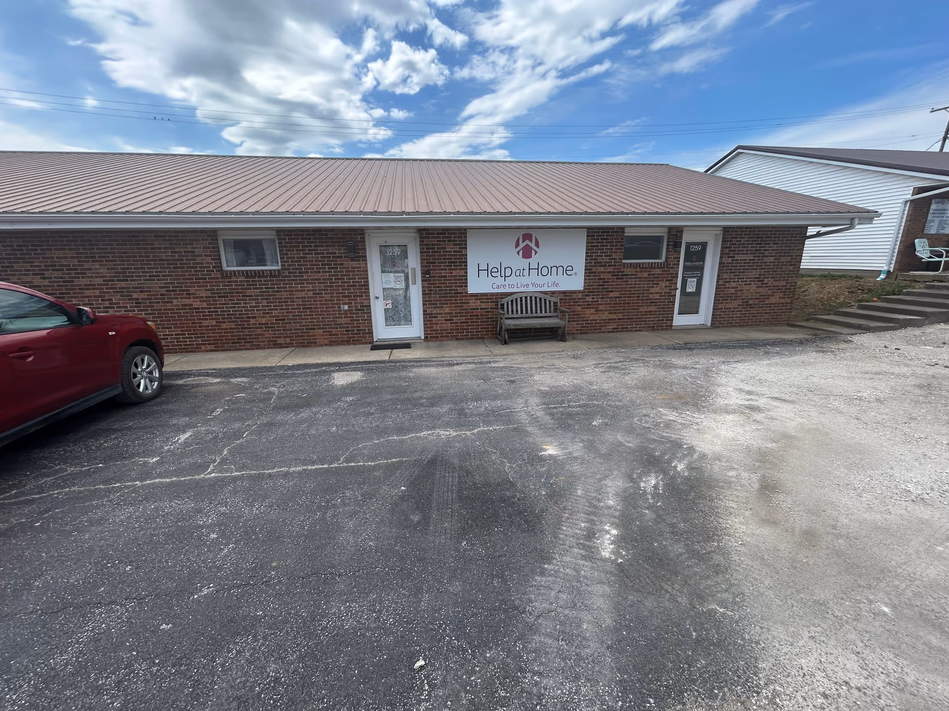 Brick exterior of Help at Home facility with front doors, banner, bench, and red car.