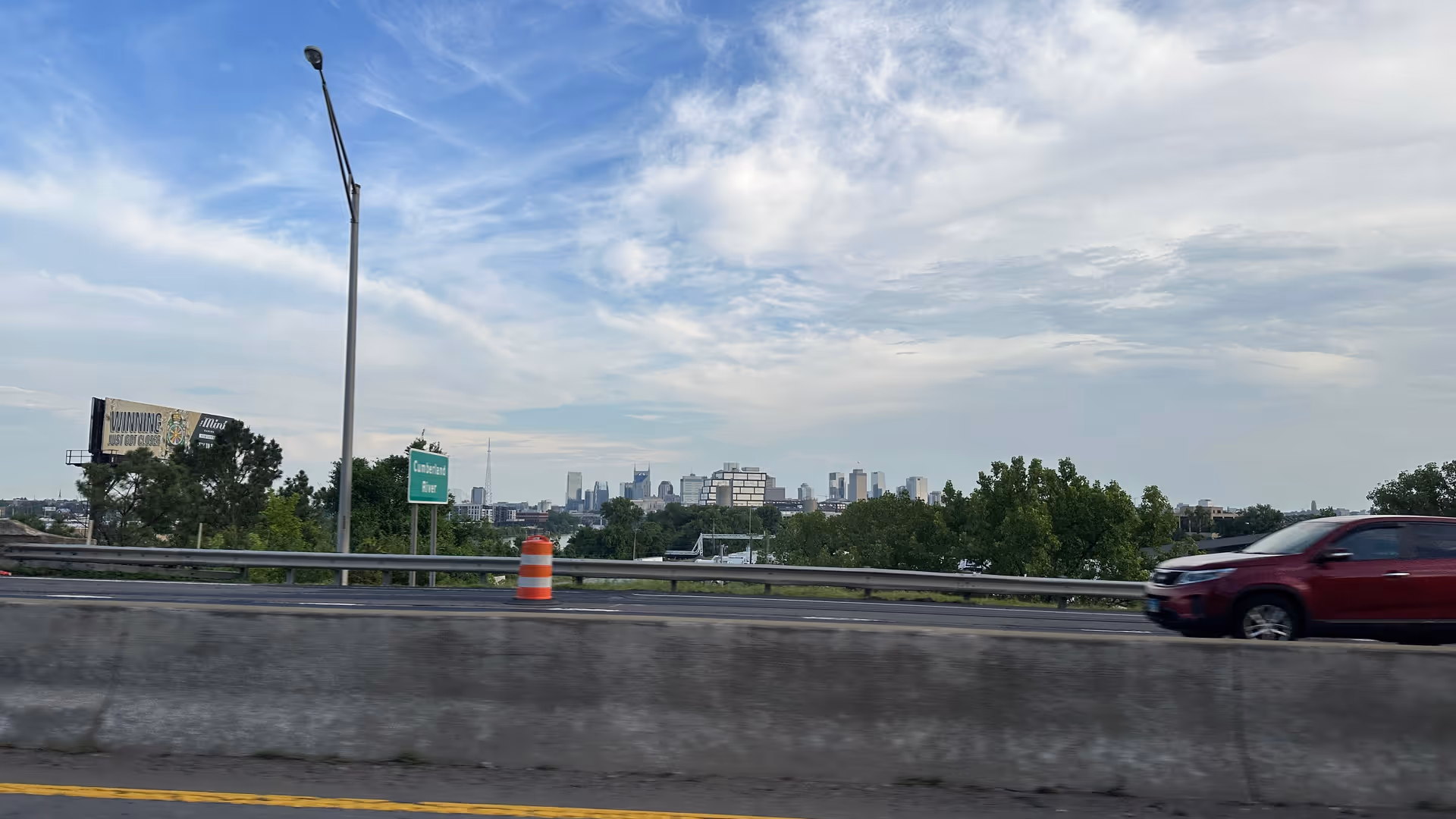 Highway scene with guardrail, orange barrel, trees, and distant city skyline.