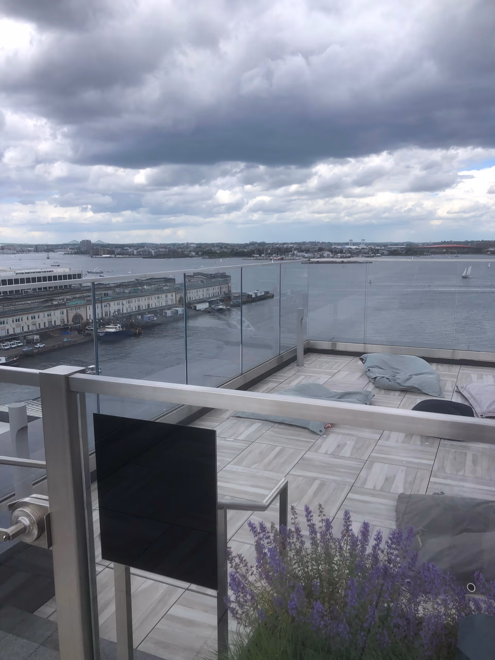 Glass-railed rooftop terrace with cushions overlooking a harbor and distant city skyline.