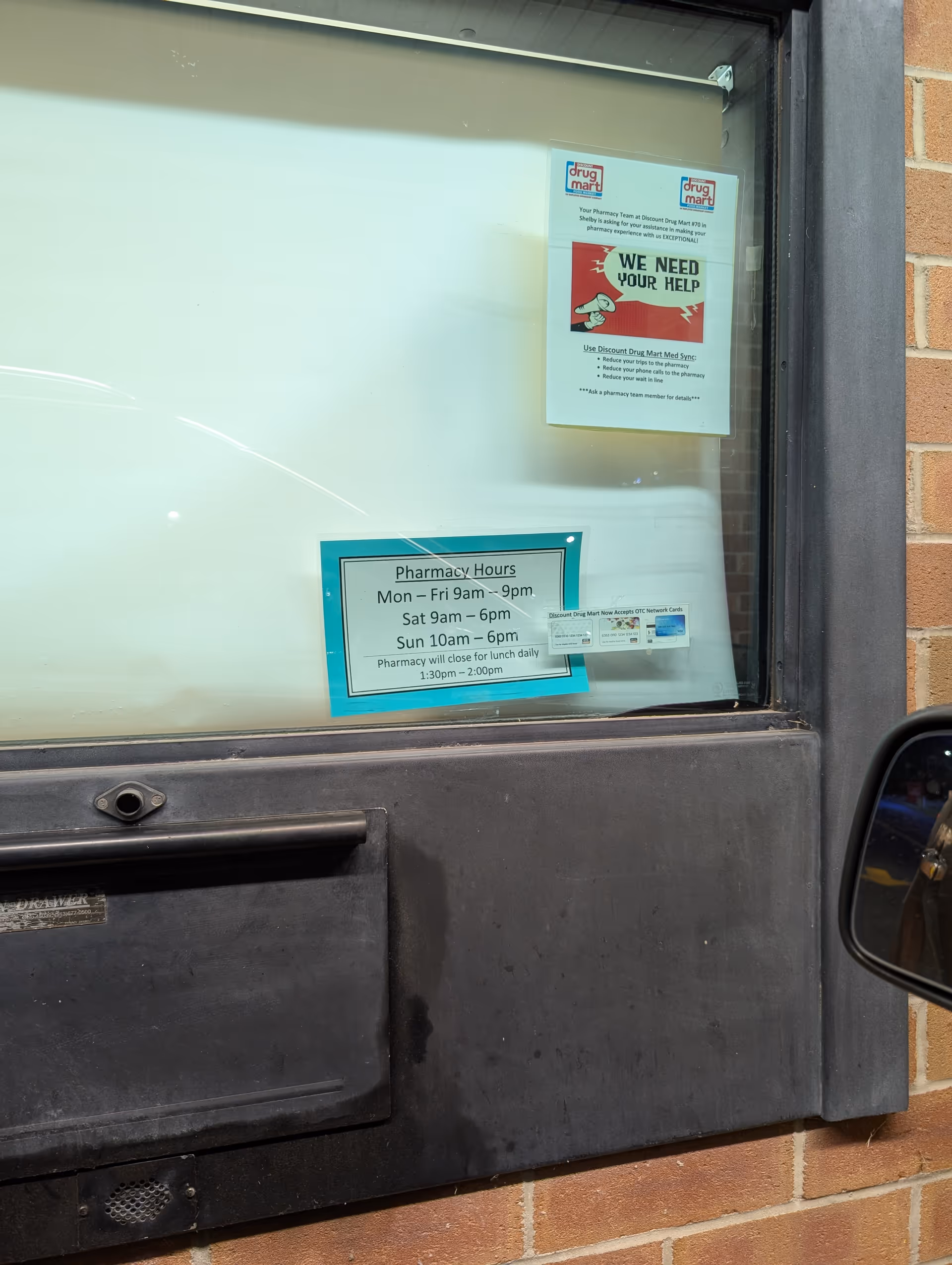 Front window of Discount Drug Mart with pharmacy hours and 'We Need Your Help' flyer.