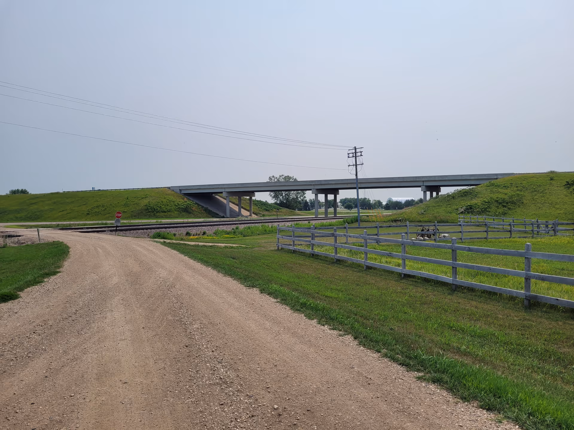 Gravel road leads to a highway overpass with grassy embankments and fences.