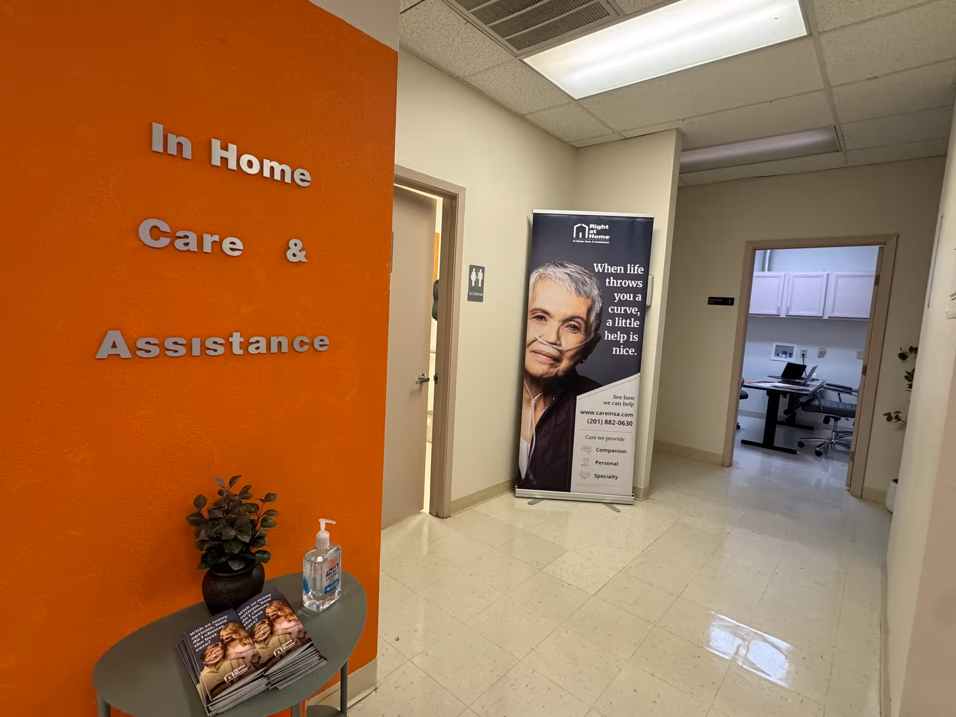 Orange wall shows 'In Home Care & Assistance' beside a hallway and brochure table.