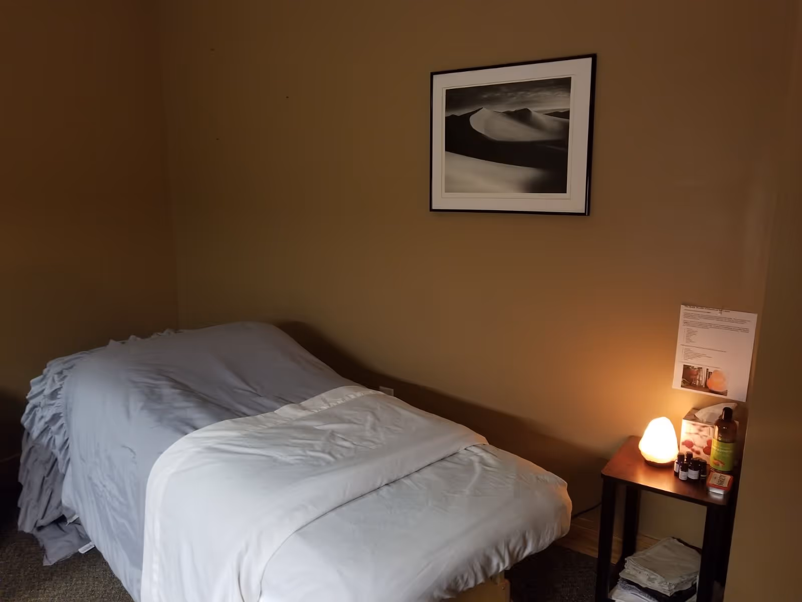 A bed with light blue sheets sits in a small beige bedroom with nightstand and lamp.