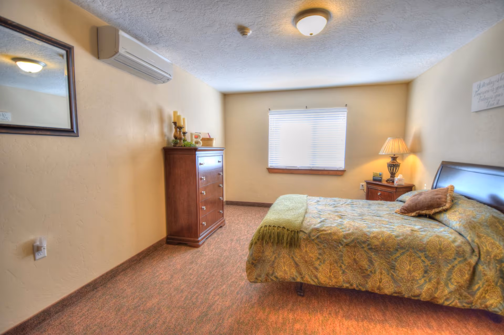A cozy bedroom features a large bed, dresser, nightstand, lamp, and window with blinds.
