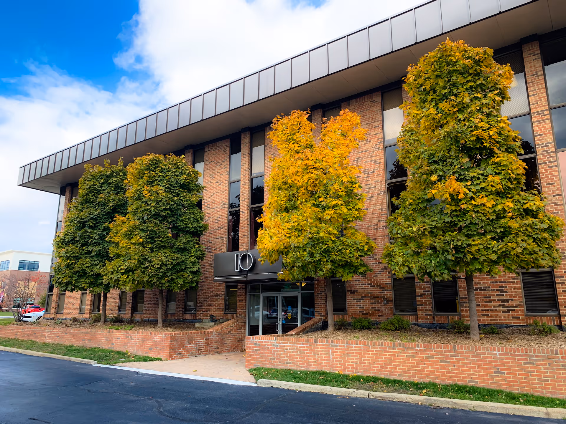 The brick senior living facility front features glass doors and three autumn-colored trees.