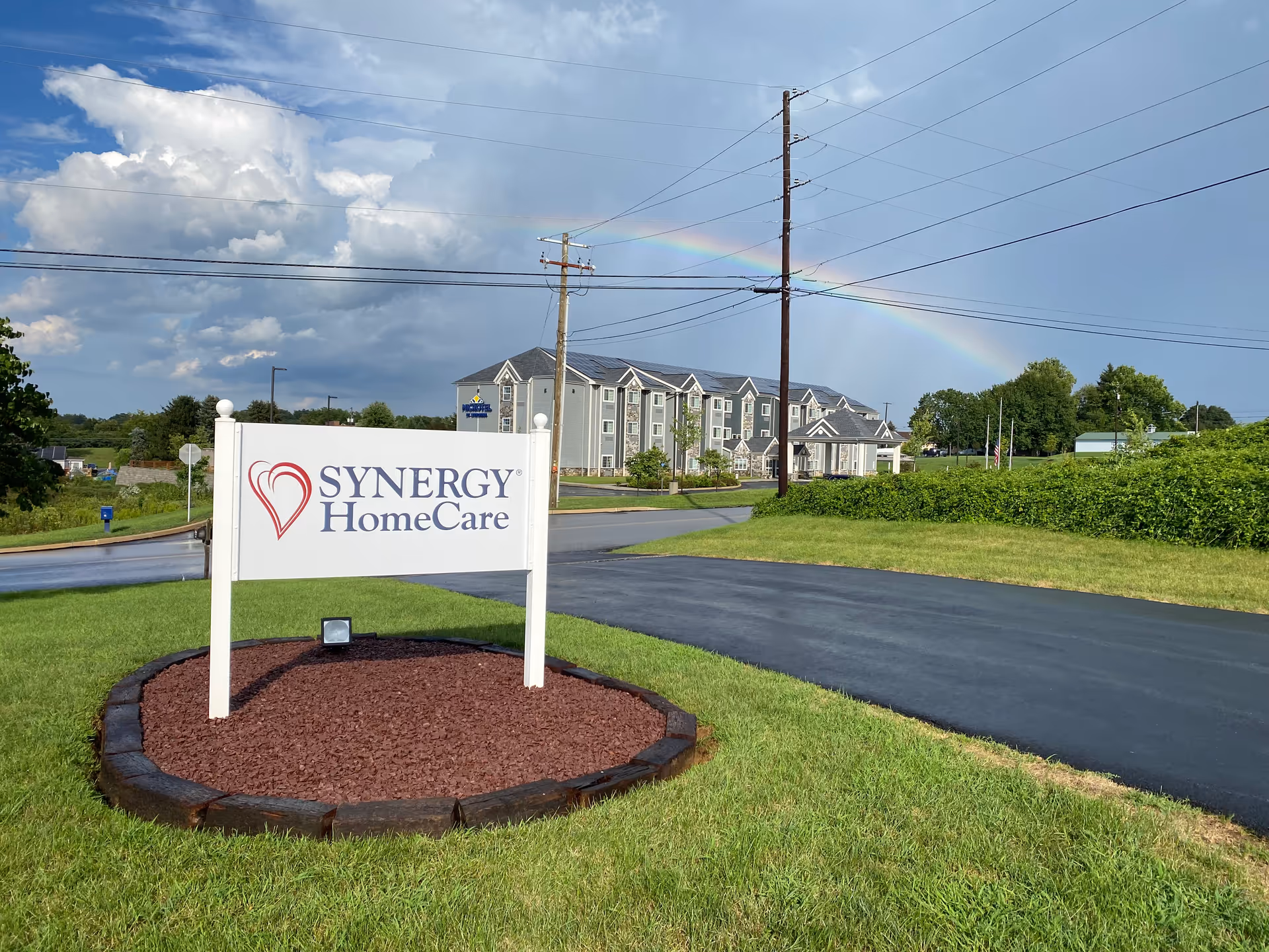 Exterior view of SYNERGY HomeCare facility with sign, road, and rainbow.