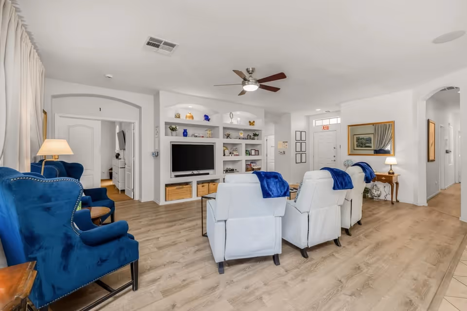 Bright communal living room with blue chairs, white recliners, TV, and built-in shelving.