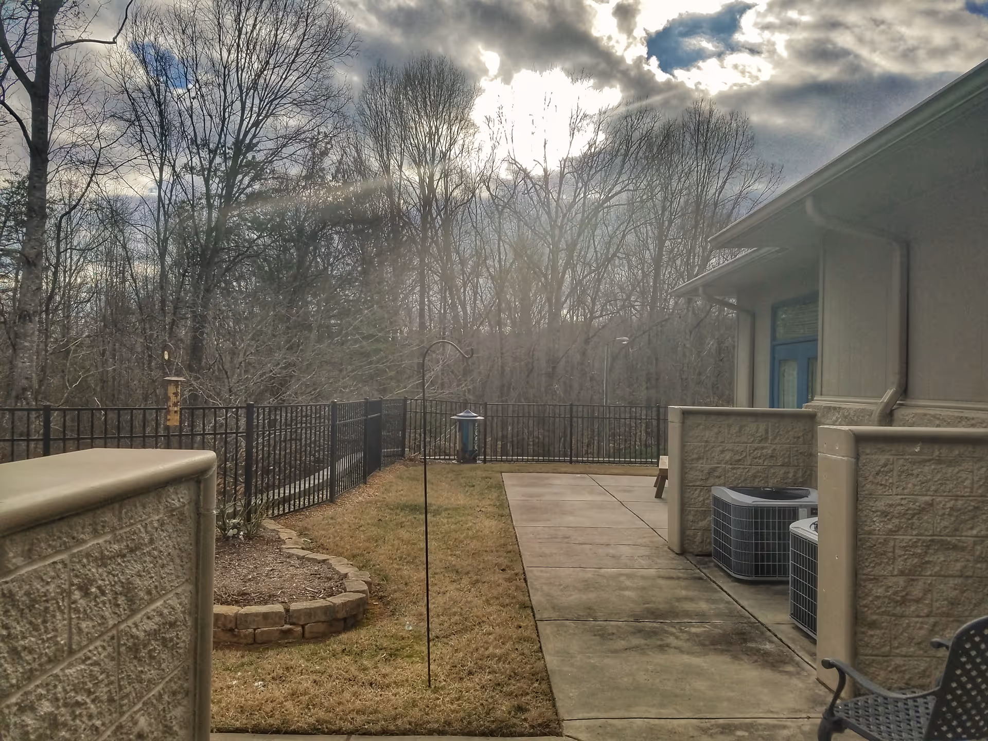 Outdoor patio at a senior living facility with trees, railing, and air conditioning units.