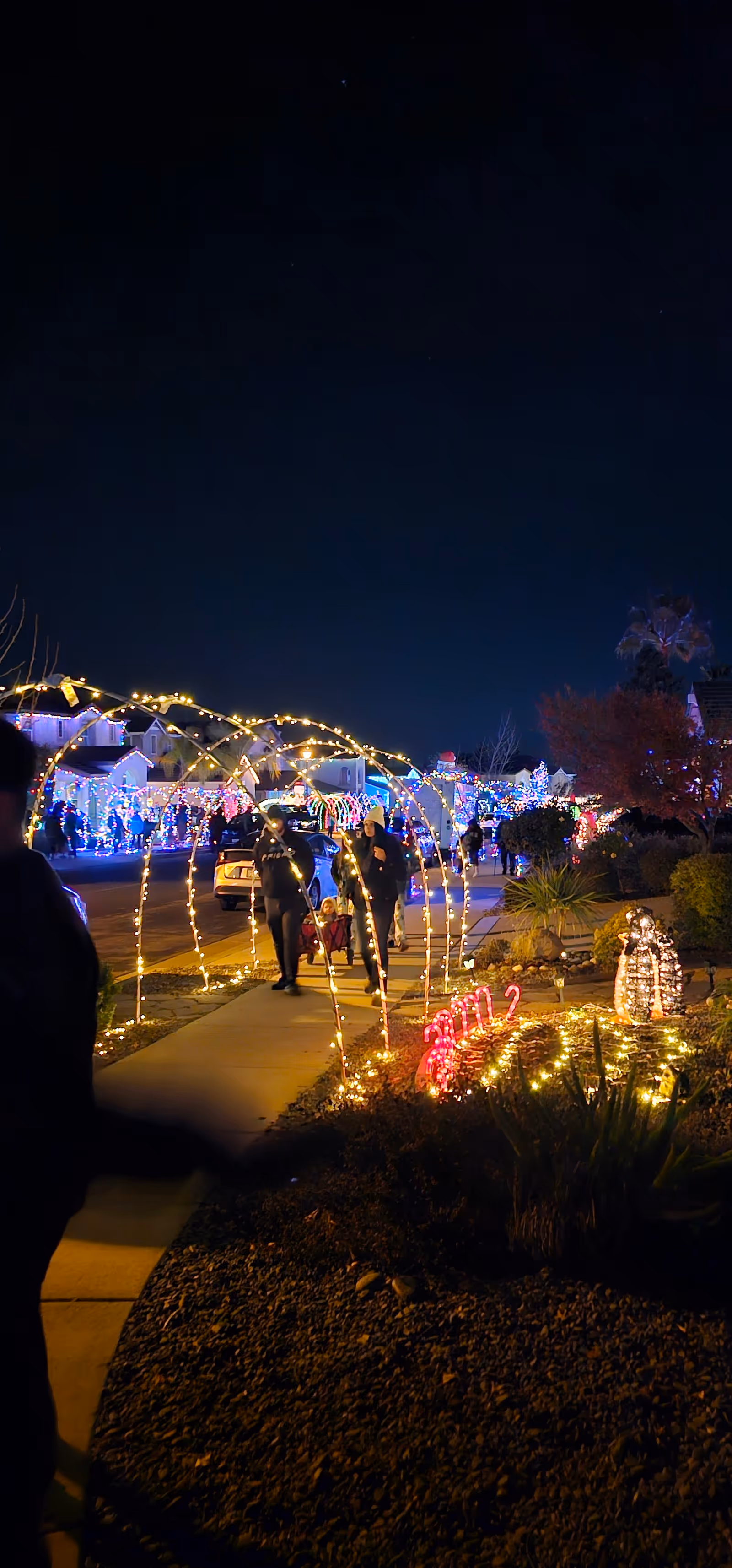 Nighttime street scene with festive lights, arches, and pedestrians along a residential neighborhood.