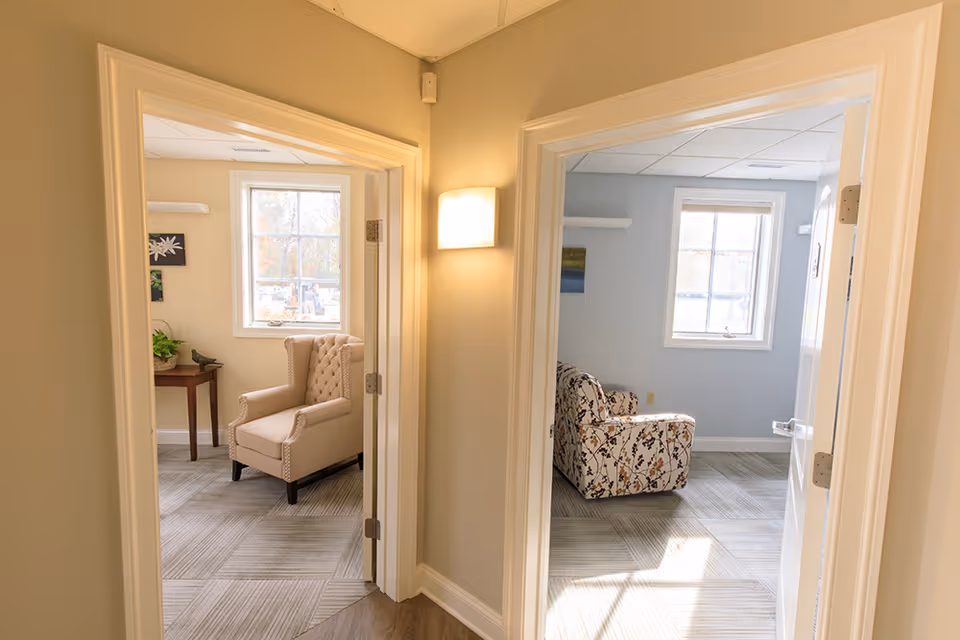 Two doorways open to separate sitting rooms with upholstered chairs in a sunlit hallway.