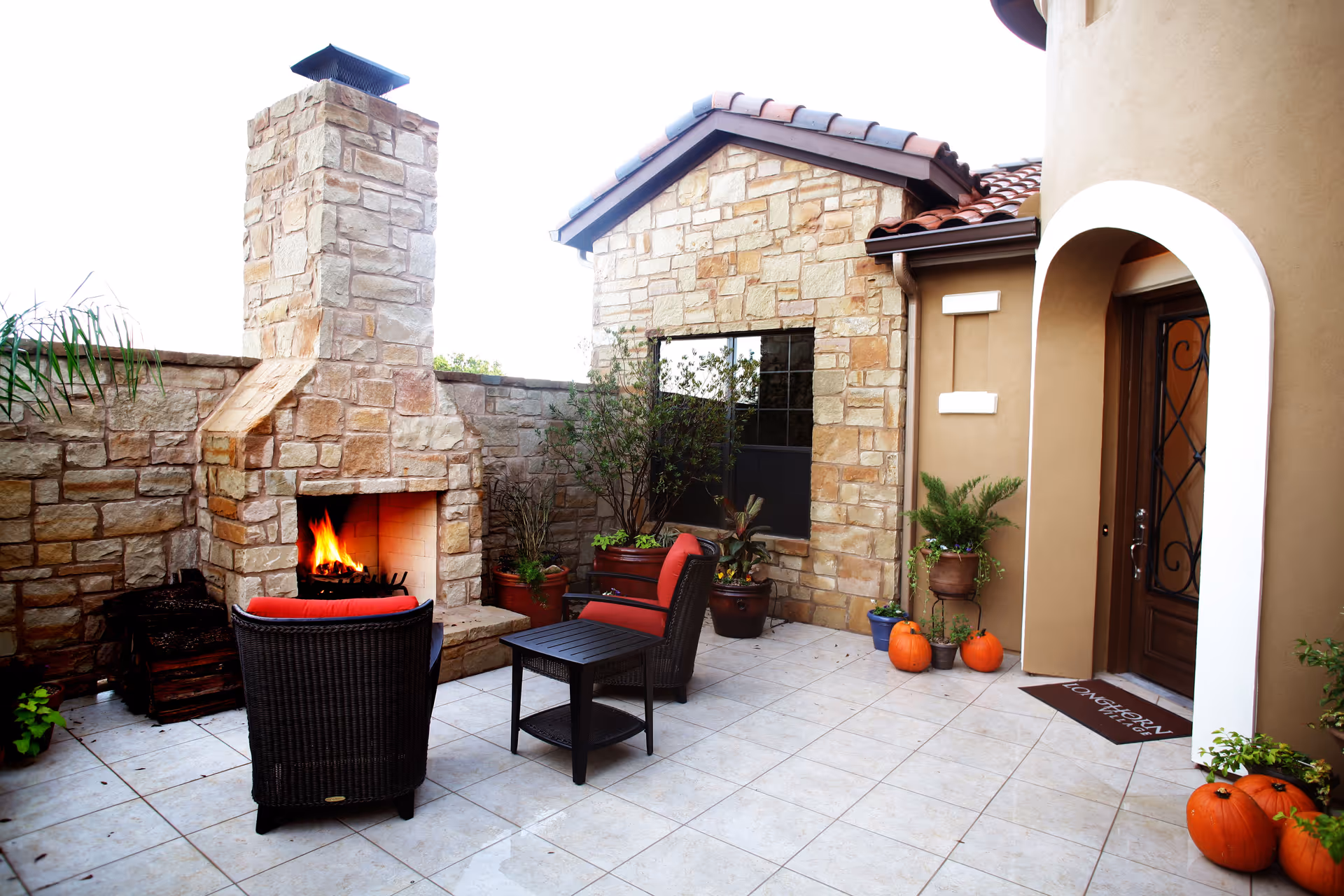 Stone-walled outdoor patio with a fireplace, seating, potted plants, pumpkins, and an arched entrance.