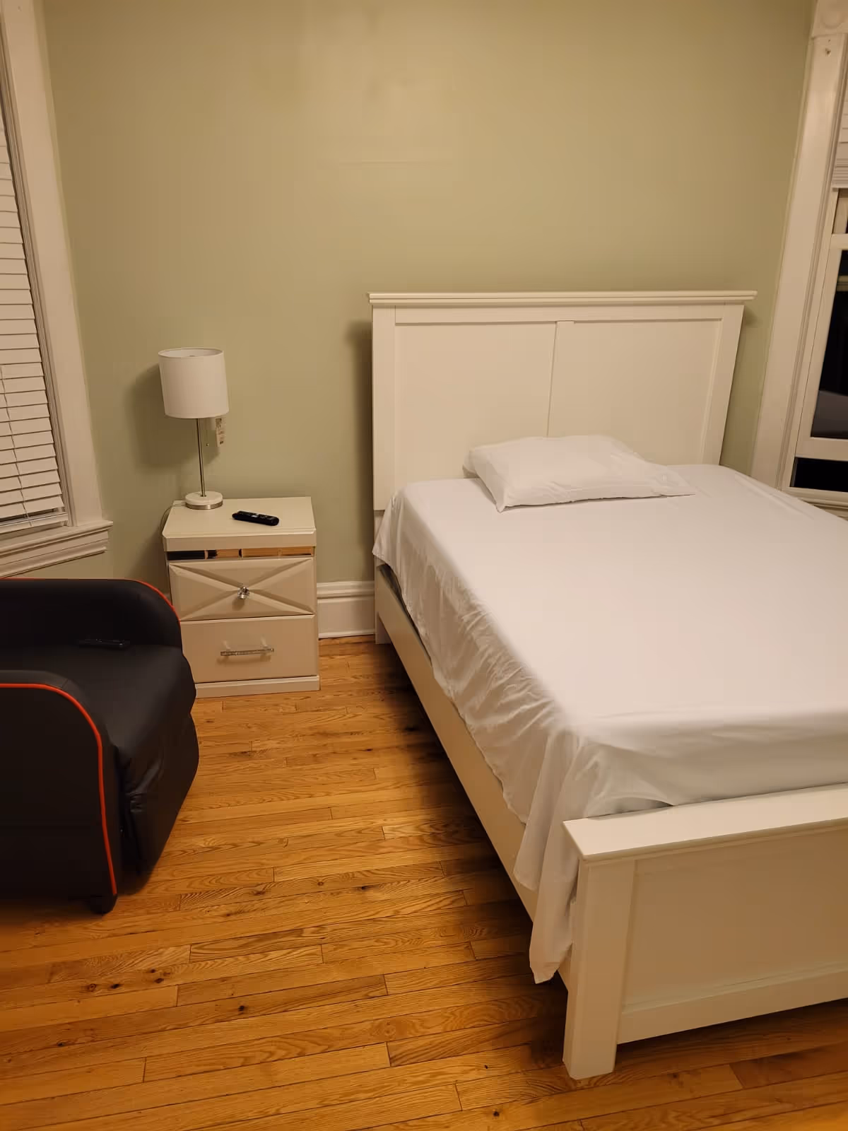White bed with pillow, nightstand lamp, and chair in a pale green bedroom.