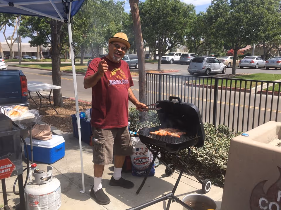 A man in a hat grills sausages on a charcoal barbecue outdoors under a canopy.