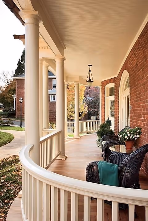 Covered wooden porch with columns, wicker chairs, and potted plants along a brick building.