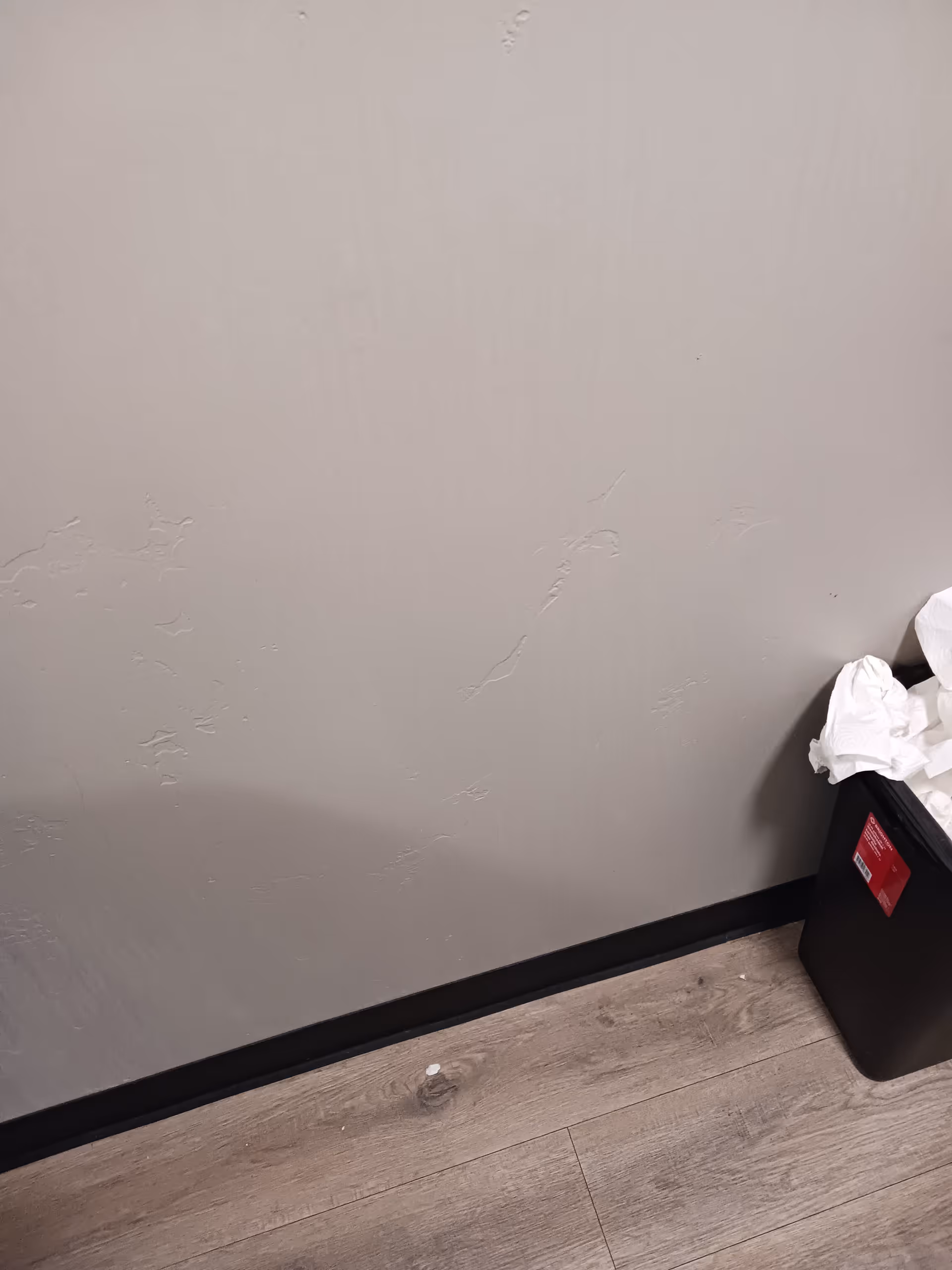 Corner of a light gray wall with a black trash bin and crumpled papers.
