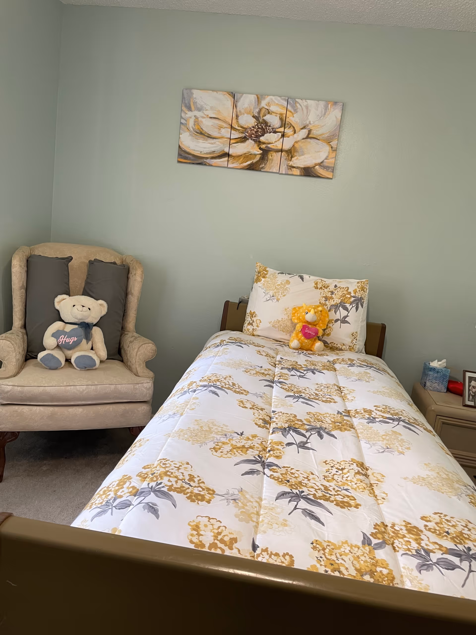 Cozy bedroom in a senior facility with a floral bedspread, chair, and stuffed bears.