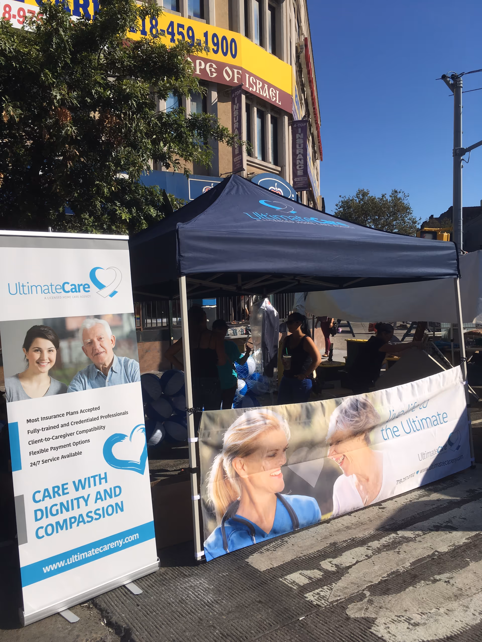 Outdoor promotional tent for Ultimate Care NY with banners and people nearby.
