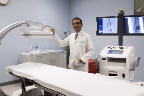 A doctor in a white coat stands beside medical imaging equipment in a clinical room.