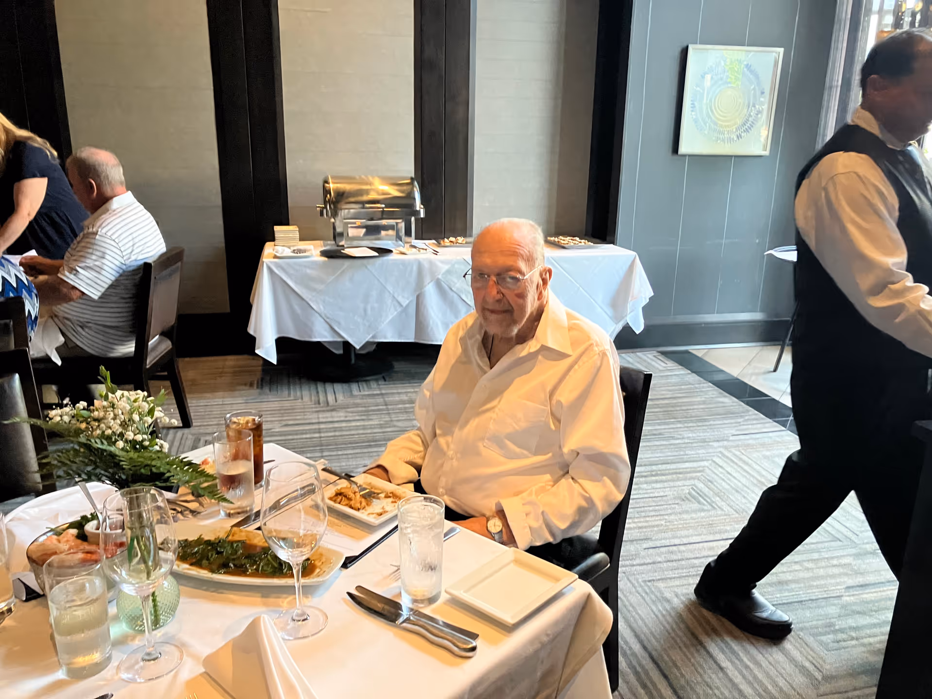 An elderly man in a white shirt sits at a dining table with meals and glassware.